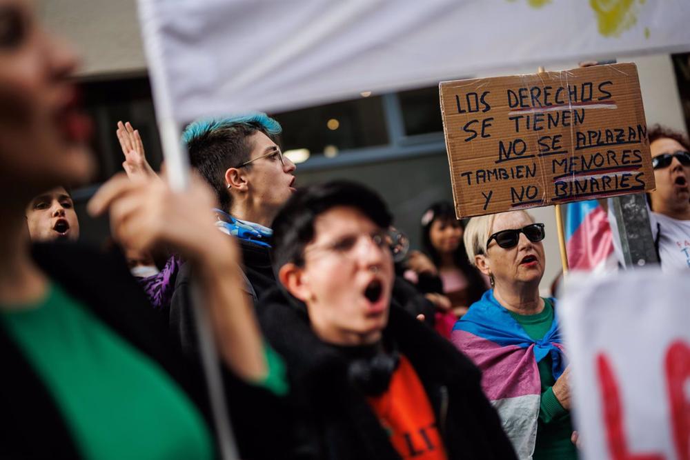 Archivo - Una mujer con una bandera trans sujeta un cartel en el que se lee: 'Los derechos se tienen no se aplazan' Archivo